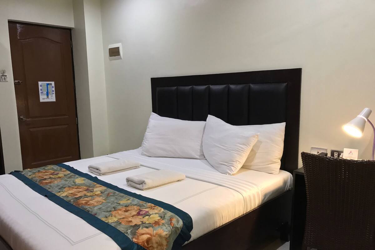 A neatly made bed with white linens and a floral-patterned bedspread, a dark headboard, and a bedside lamp.
