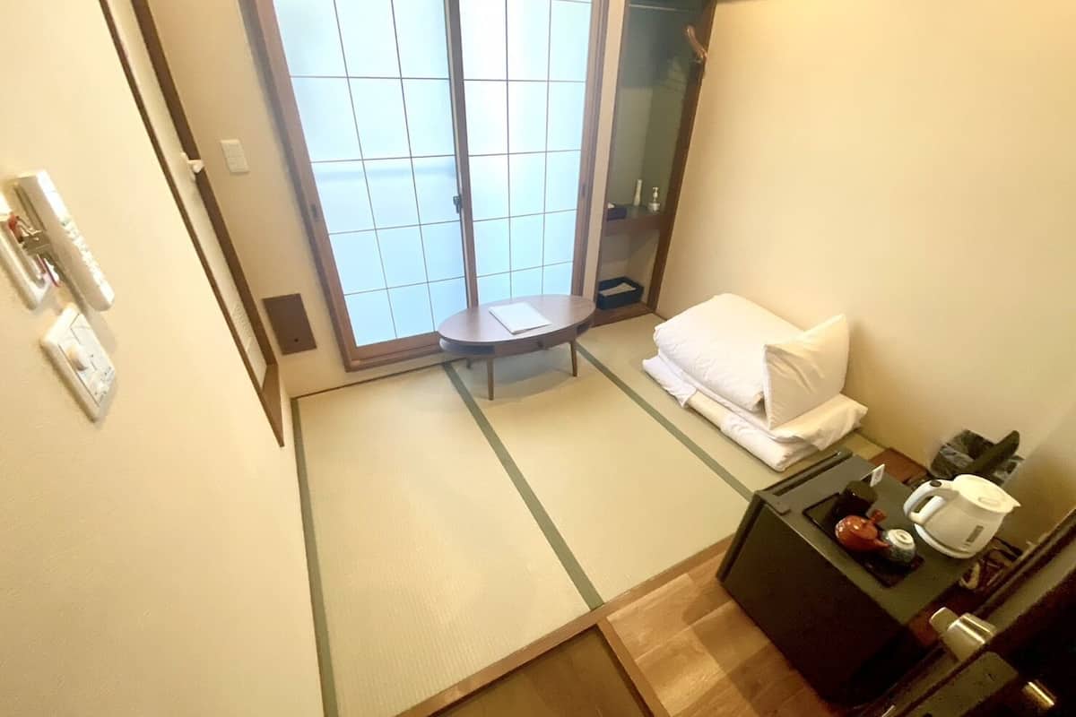 Quarto de hotel com porta deslizante para uma varanda, uma pequena mesa redonda, uma cama com travesseiro branco e um minibar.