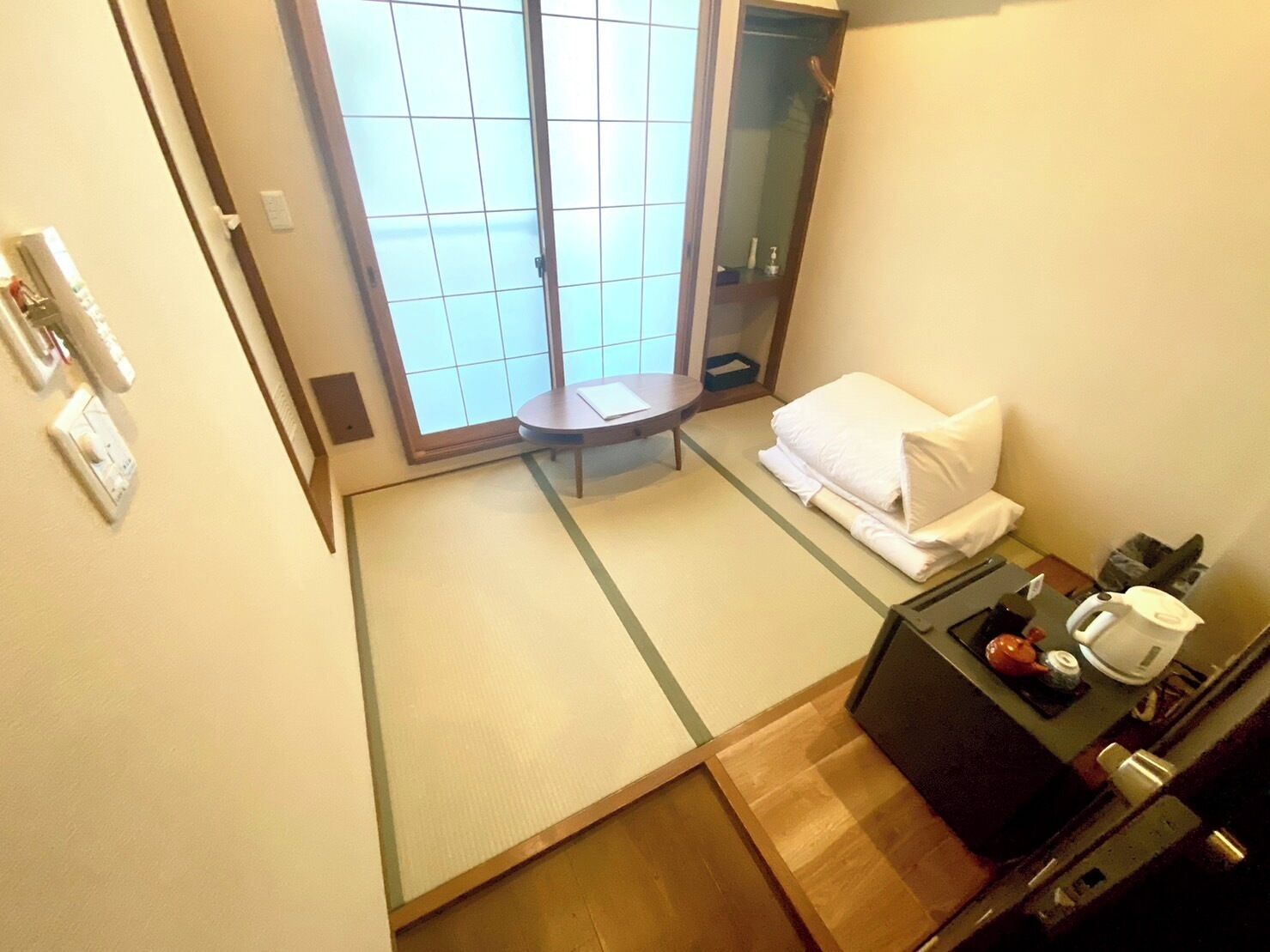 Habitación de hotel con puerta corredera, una pequeña mesa con una silla, una cama con almohada y manta blancos, una mesita de noche con hervidor y taza, y un espejo.