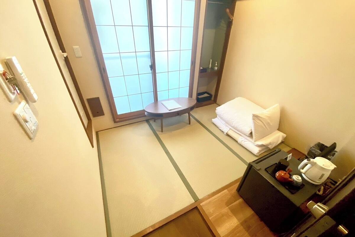 A hotel room with a sliding door, a small table with a chair, a bed with a white pillow and blanket, a bedside table with a kettle and a cup, and a mirror.