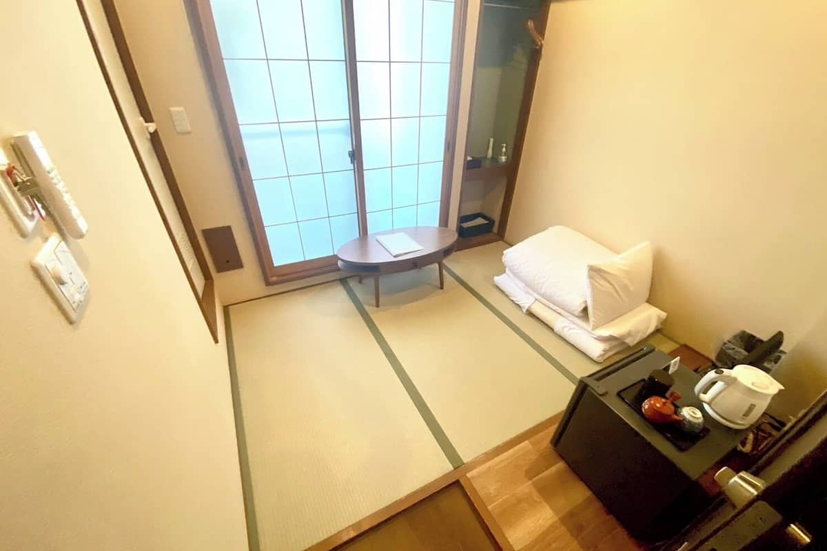 Habitación de hotel con puerta corredera, una pequeña mesa con una silla, una cama con almohada y manta blancos, una mesita de noche con hervidor y taza, y un espejo.