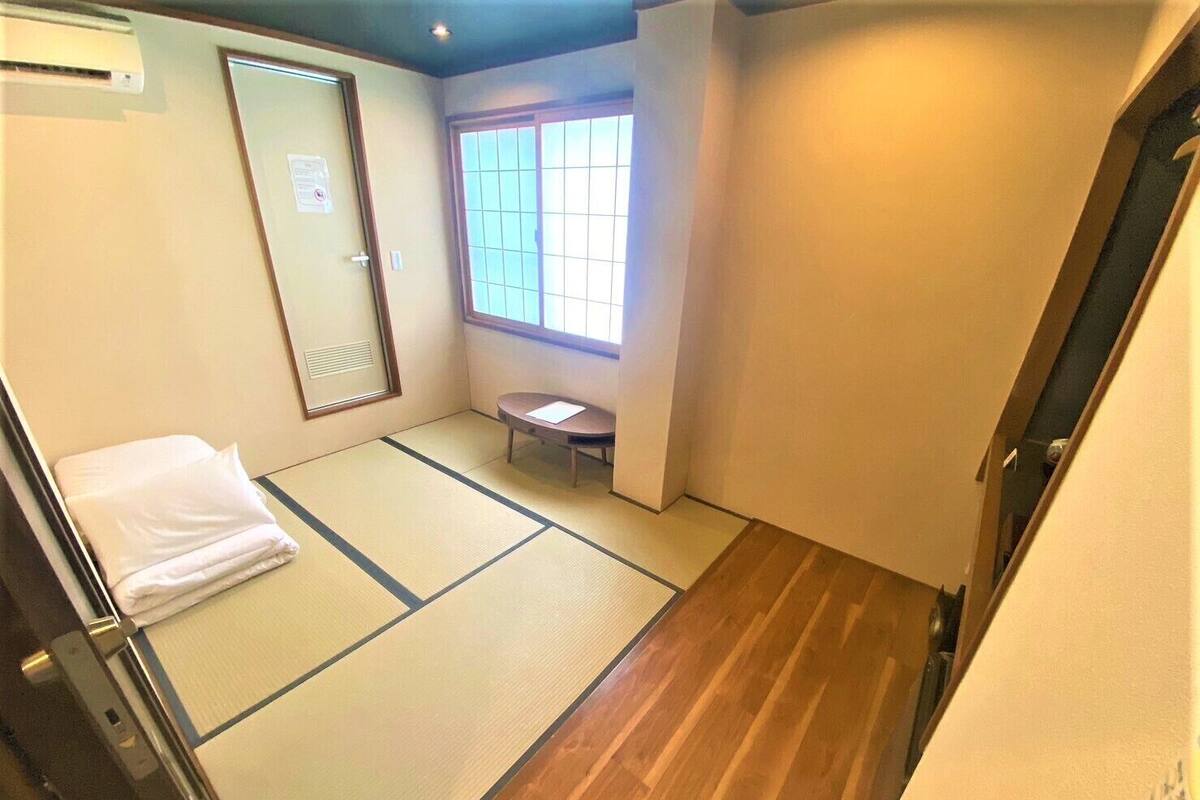 A room with tatami flooring, a wooden floor, a window with a view, a bed with a white pillow, and a small table.
