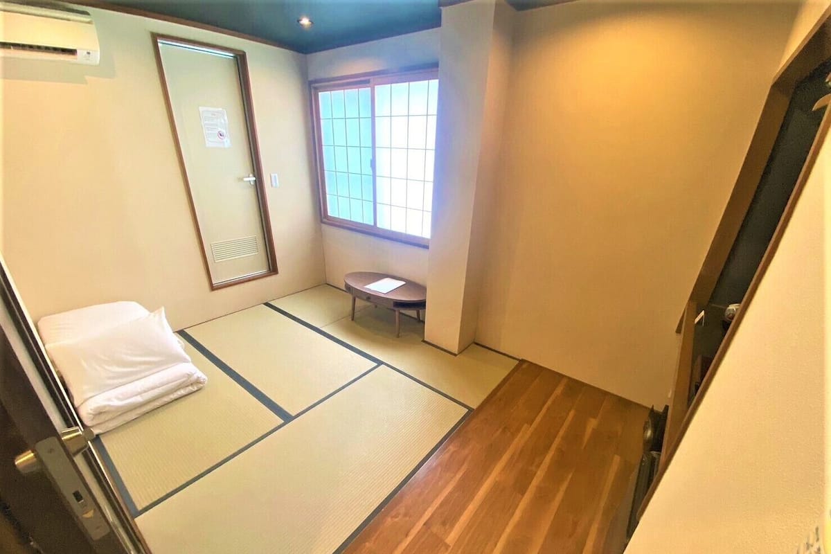 Una habitación con suelo de tatami, suelo de madera, ventana con vistas, una cama con almohada blanca y una mesita.