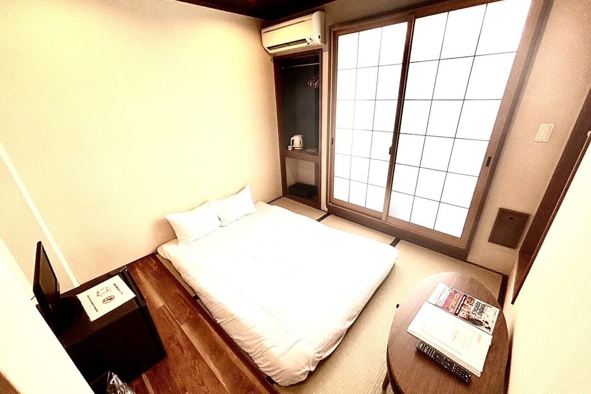 Quarto de hotel com cama, uma mesinha com revistas, um guarda-roupa e uma janela com vista.
