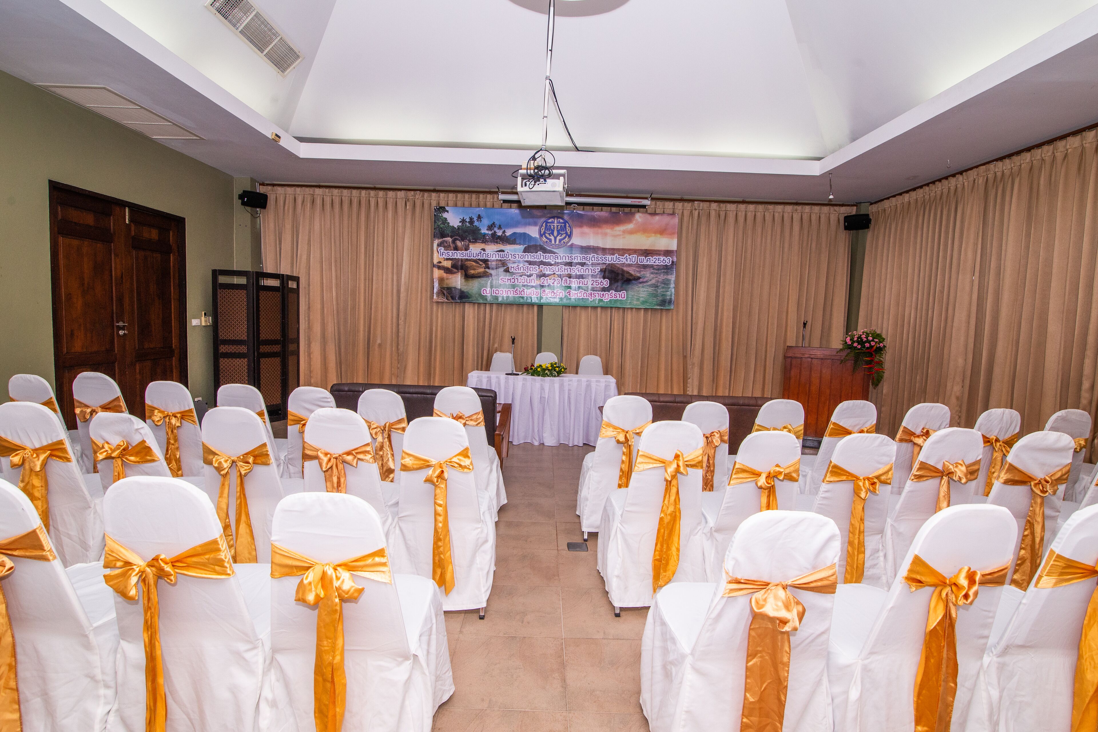 A conference room with rows of white chairs with gold sashes, a podium, a screen, and a floral arrangement.