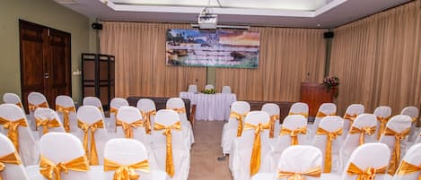 A conference room with rows of white chairs with gold sashes, a podium, a screen, and a floral arrangement.