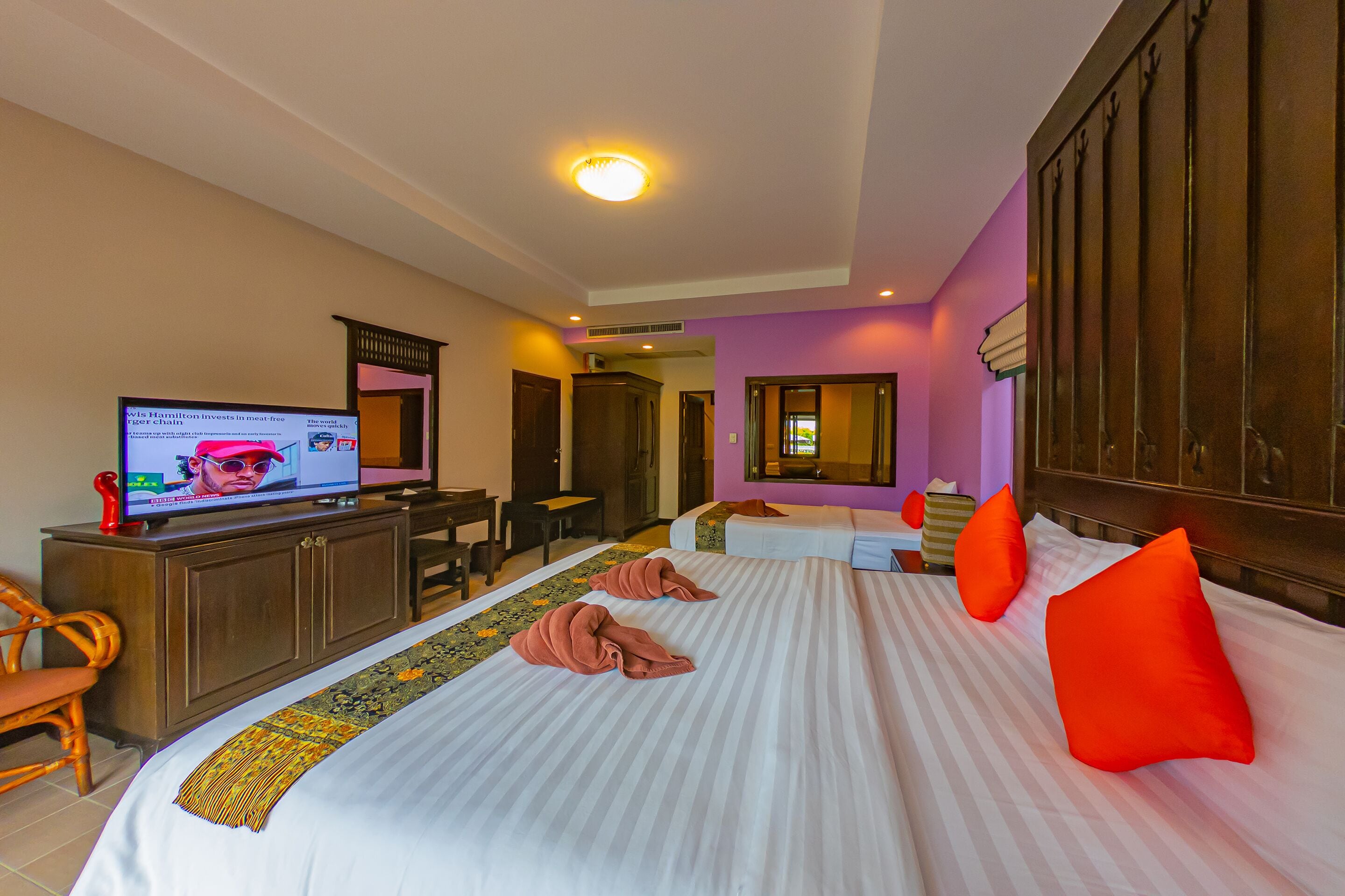 A hotel room with a television on a wooden cabinet, a bed with striped bedding and two red pillows, a wooden wardrobe, and a small desk with a chair.