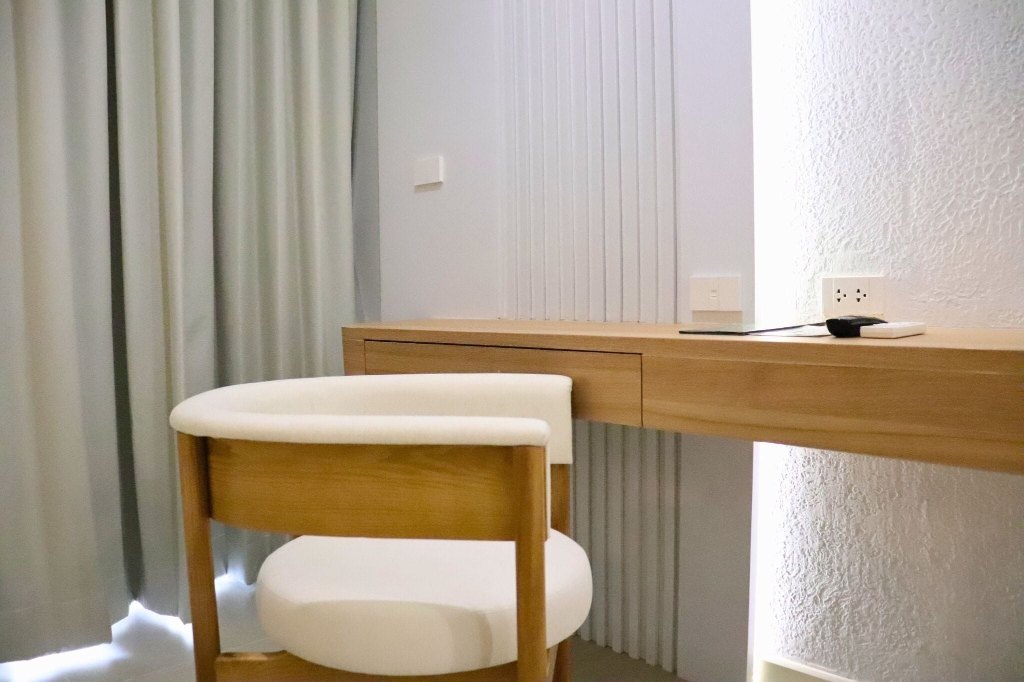 Un bureau en bois, une chaise blanche, une prise de courant et une fenêtre avec des stores.