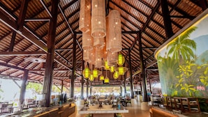 Un restaurante interior con techo alto, vigas de madera, faroles colgantes y un mural de plantas tropicales.
