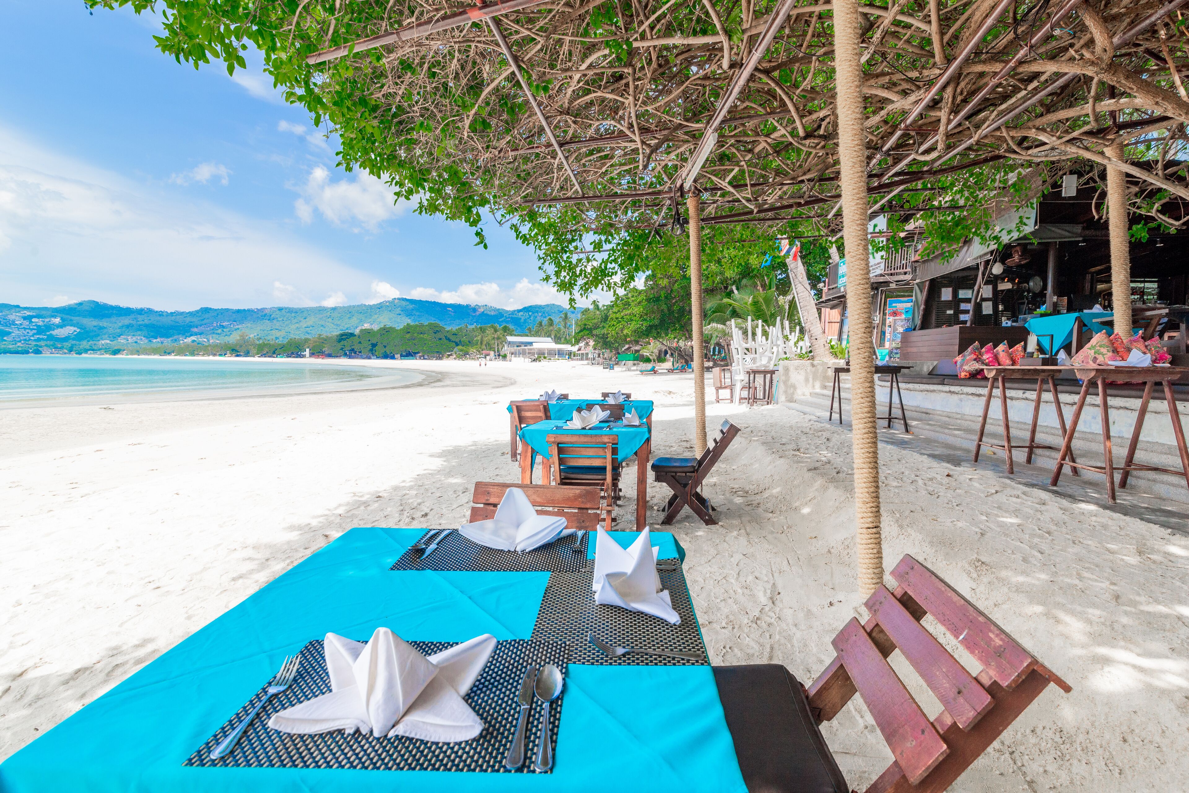 Un restaurant situé en bord de mer, avec une terrasse comprenant des tables et des chaises, offrant une vue sur une plage de sable et l’océan.