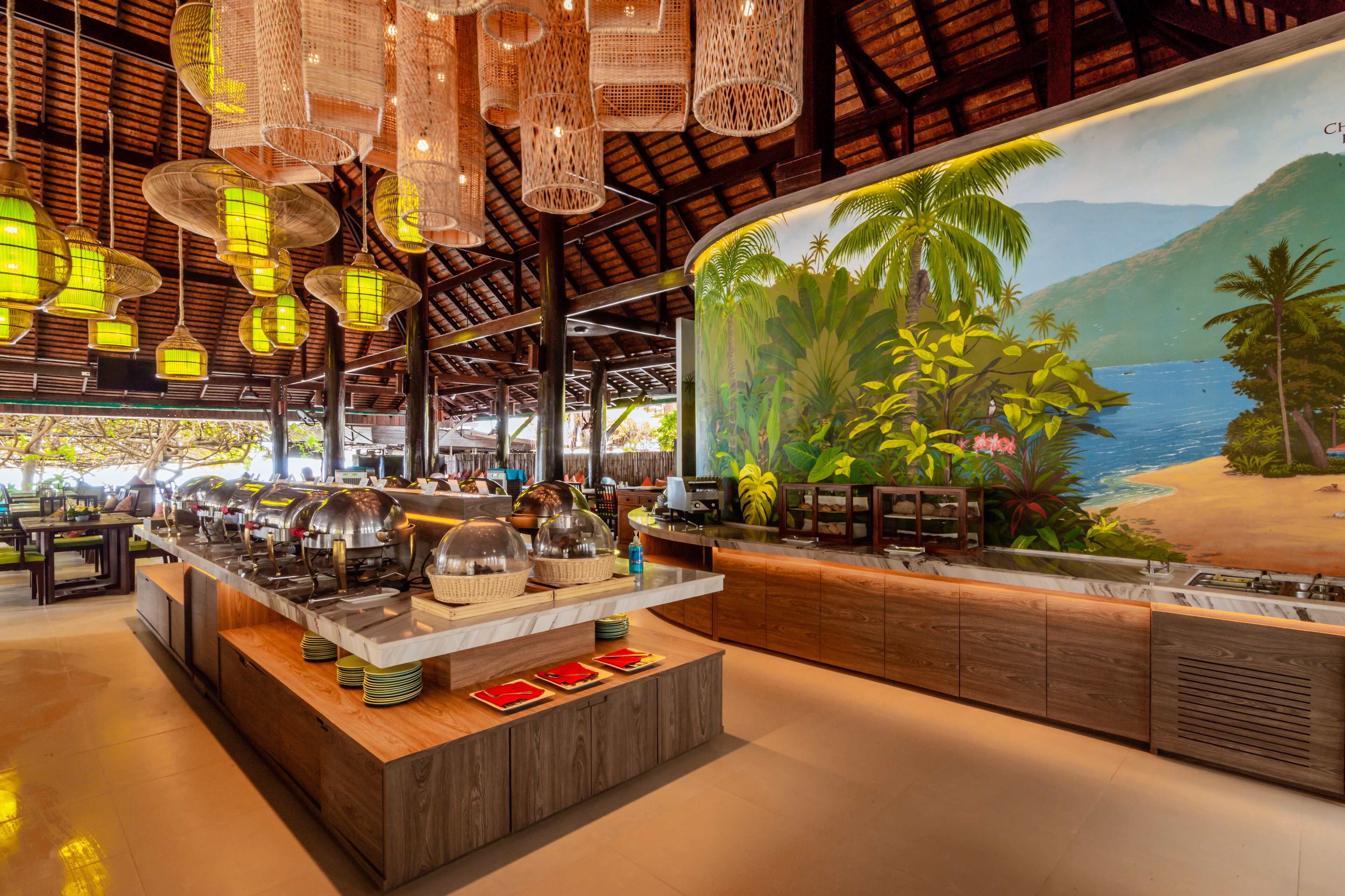 A buffet setup with a tropical mural, hanging lanterns, and wooden furniture.