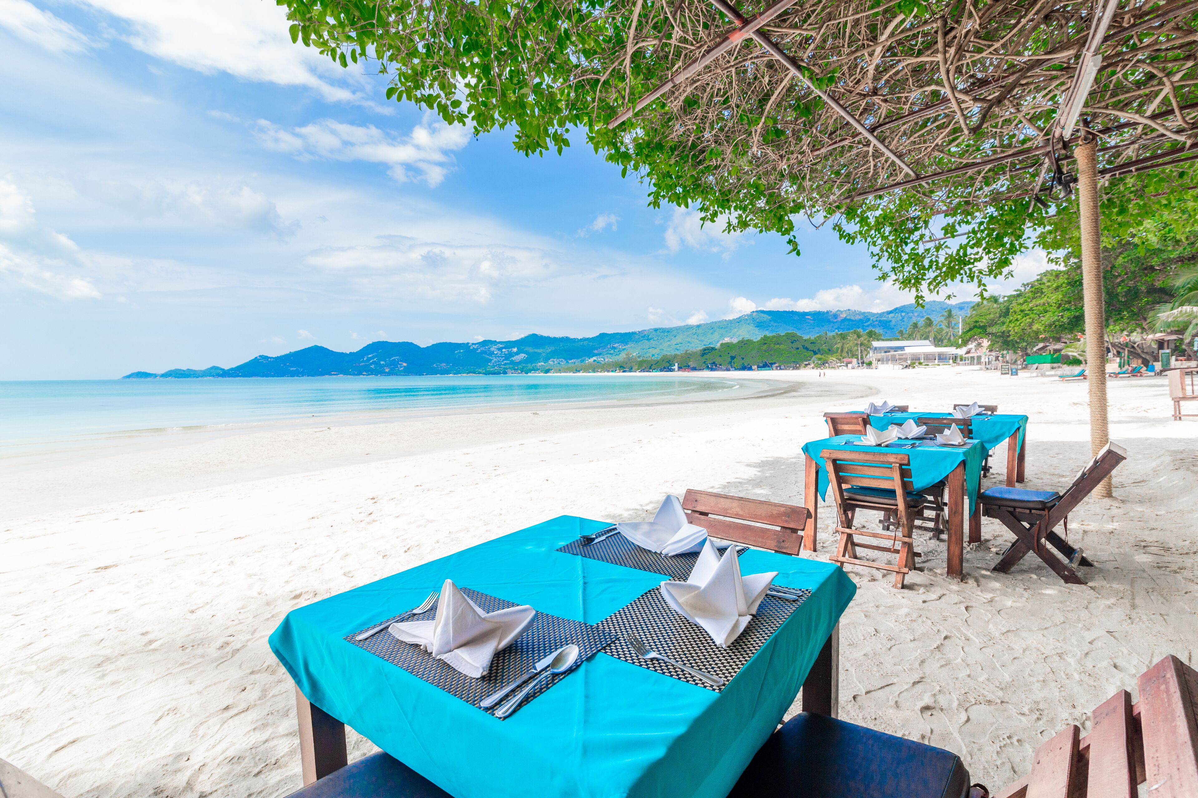 A beachside restaurant with tables set for dining, overlooking a sandy beach and the ocean.