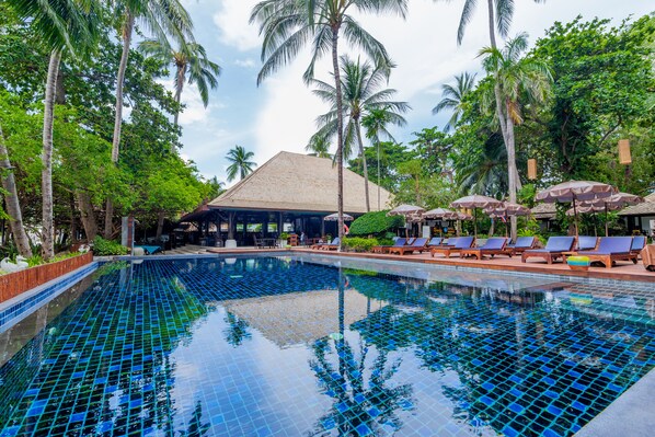 A swimming pool with tiled blue and green patterns, surrounded by palm trees and lounge chairs with umbrellas.