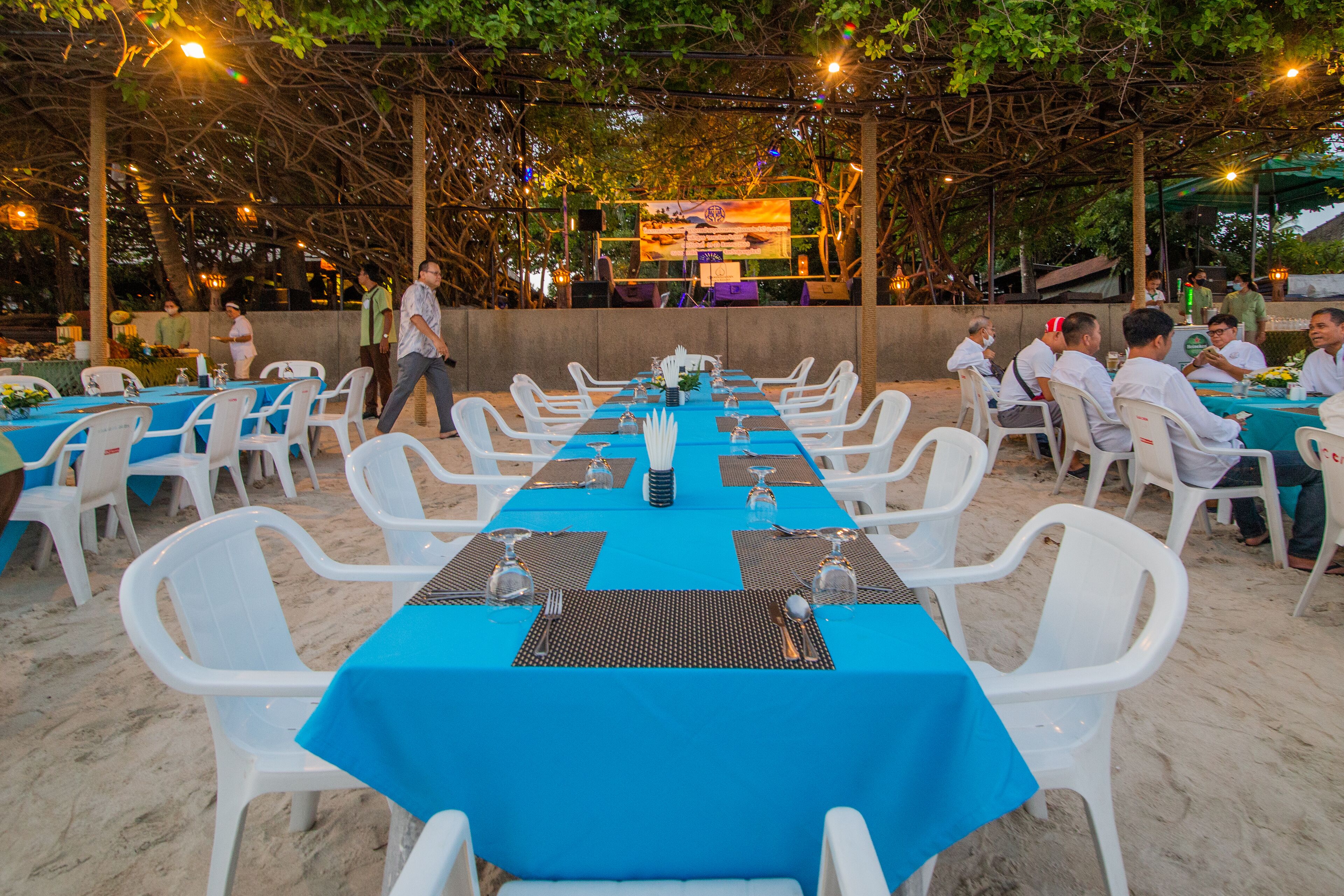 An outdoor dining area with tables set for multiple guests, surrounded by white plastic chairs, under a covered structure with string lights.