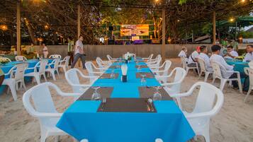 An outdoor dining area with tables set for multiple guests, surrounded by white plastic chairs, under a covered structure with string lights.