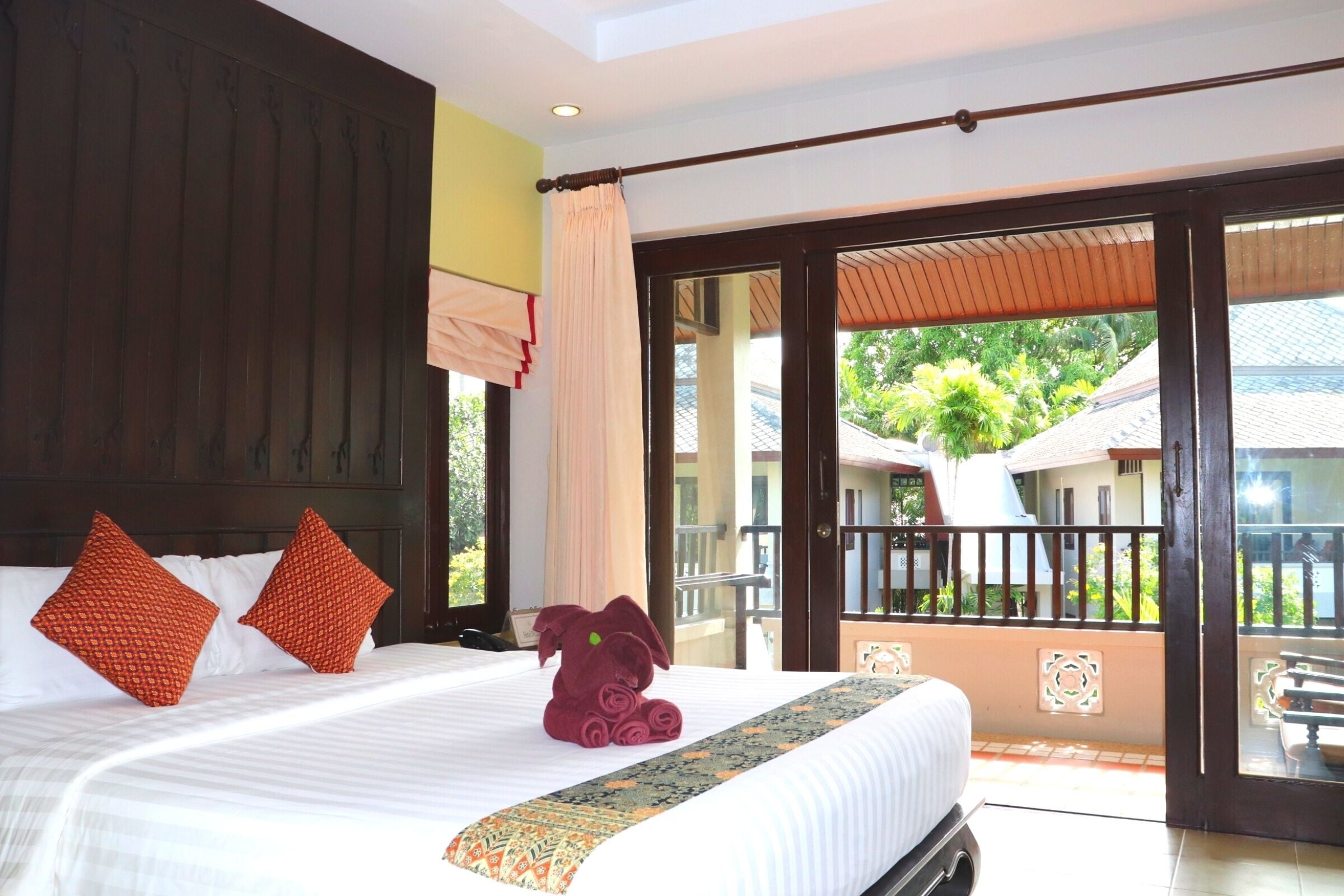 A hotel room with a large bed, two red pillows, a rolled towel, and a view of the outside balcony.