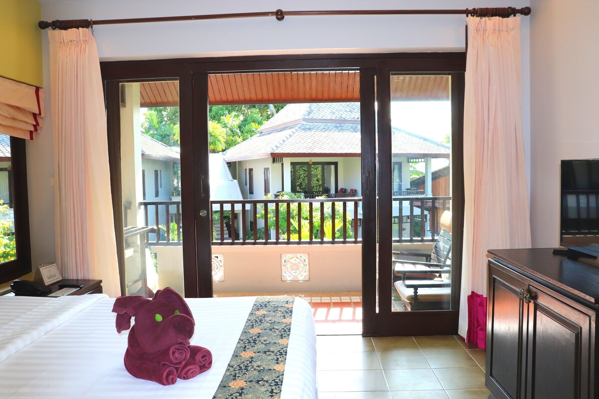 Une chambre d’hôtel avec un lit, un jouet en peluche, une télévision et un balcon donnant sur une maison et un espace verdoyant.