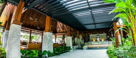A covered outdoor area with a tiled floor, concrete pillars, and lush greenery.