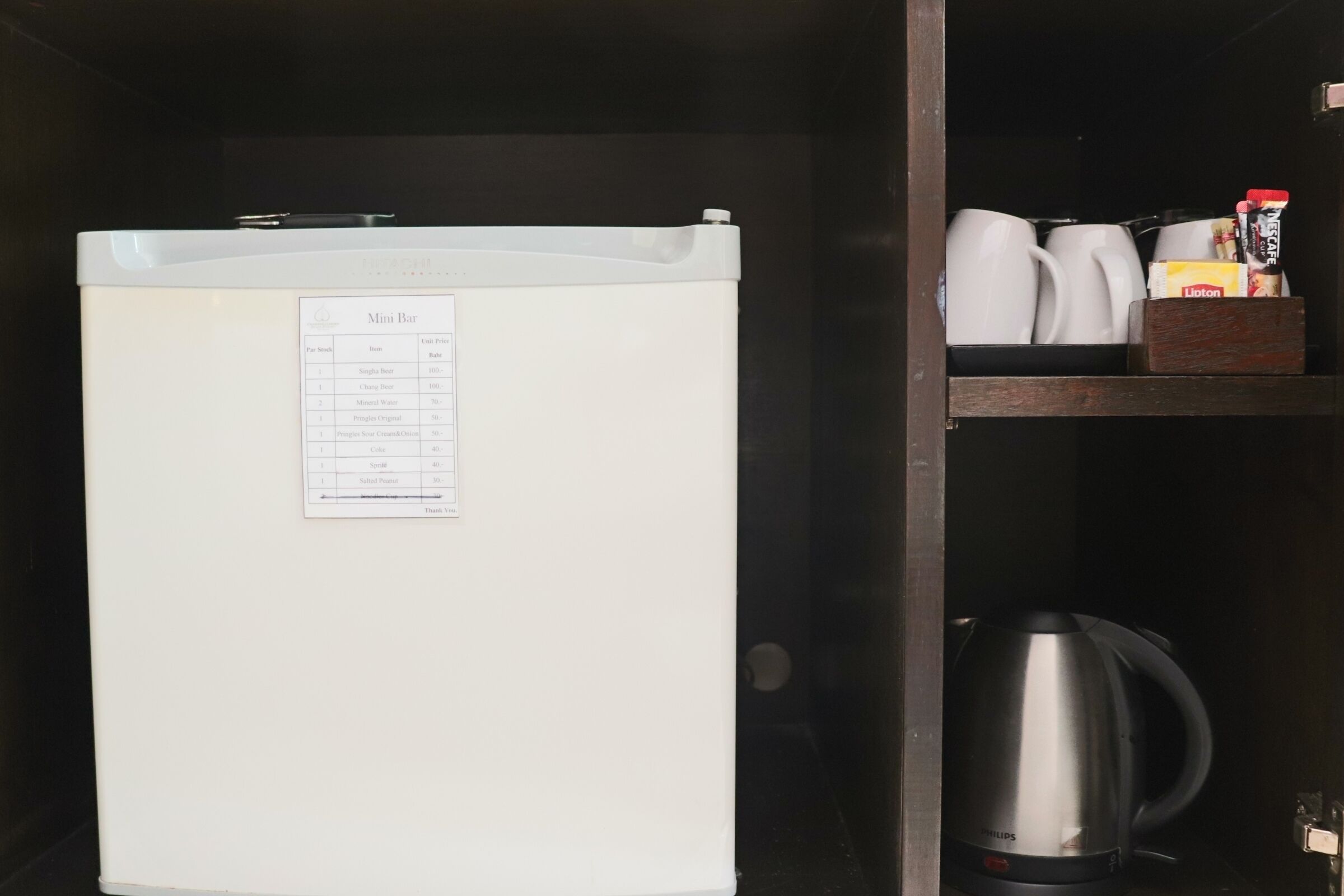 A mini fridge with a menu listing items and prices, a coffee maker, and a shelf with mugs and a box of Lipton tea.
