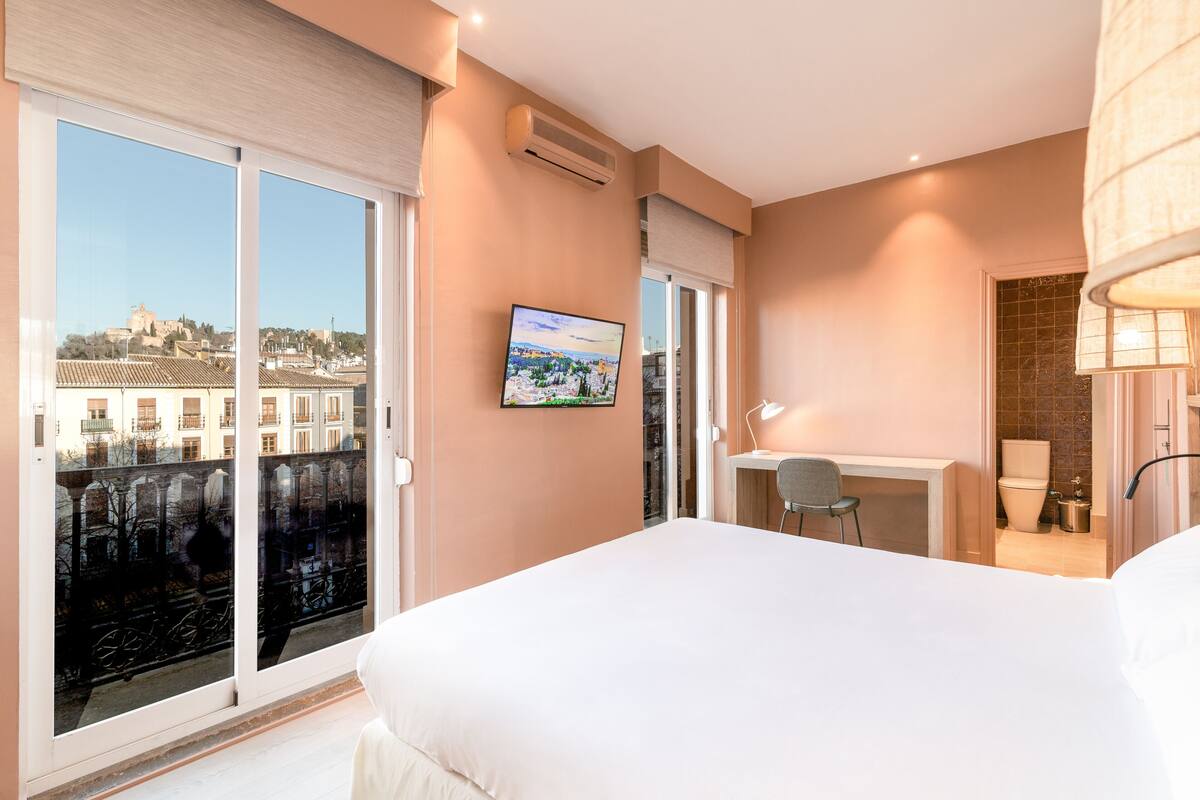 A hotel room with a bed, a desk, a chair, a TV, and a view of the city through large windows.