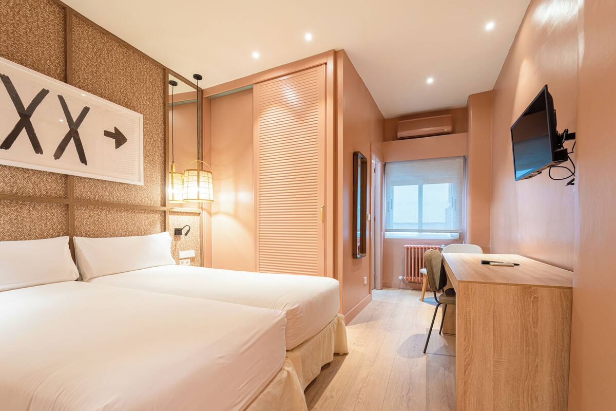 A modern hotel room with a large bed, a desk with a chair, a TV mounted on the wall, and a window with blinds.