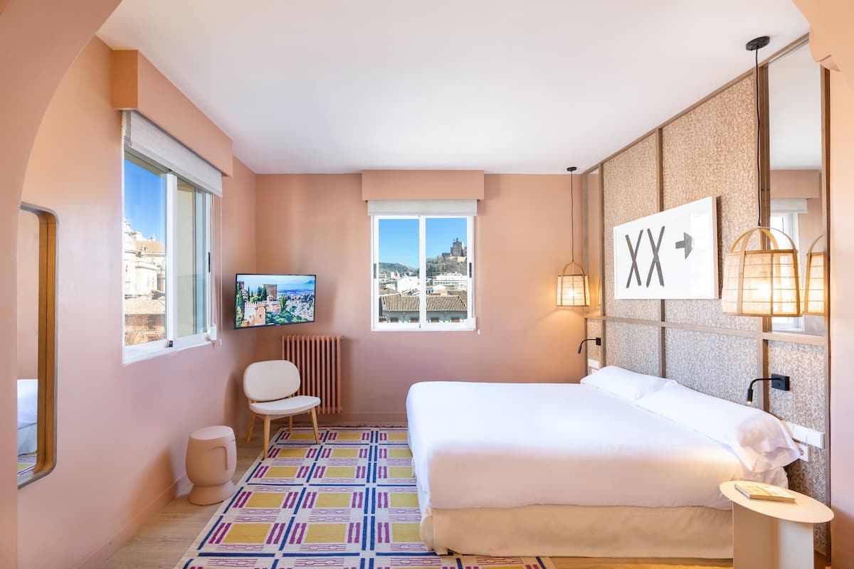 A hotel room with a bed, a TV, a chair, a small table, and a view of the city through the windows.