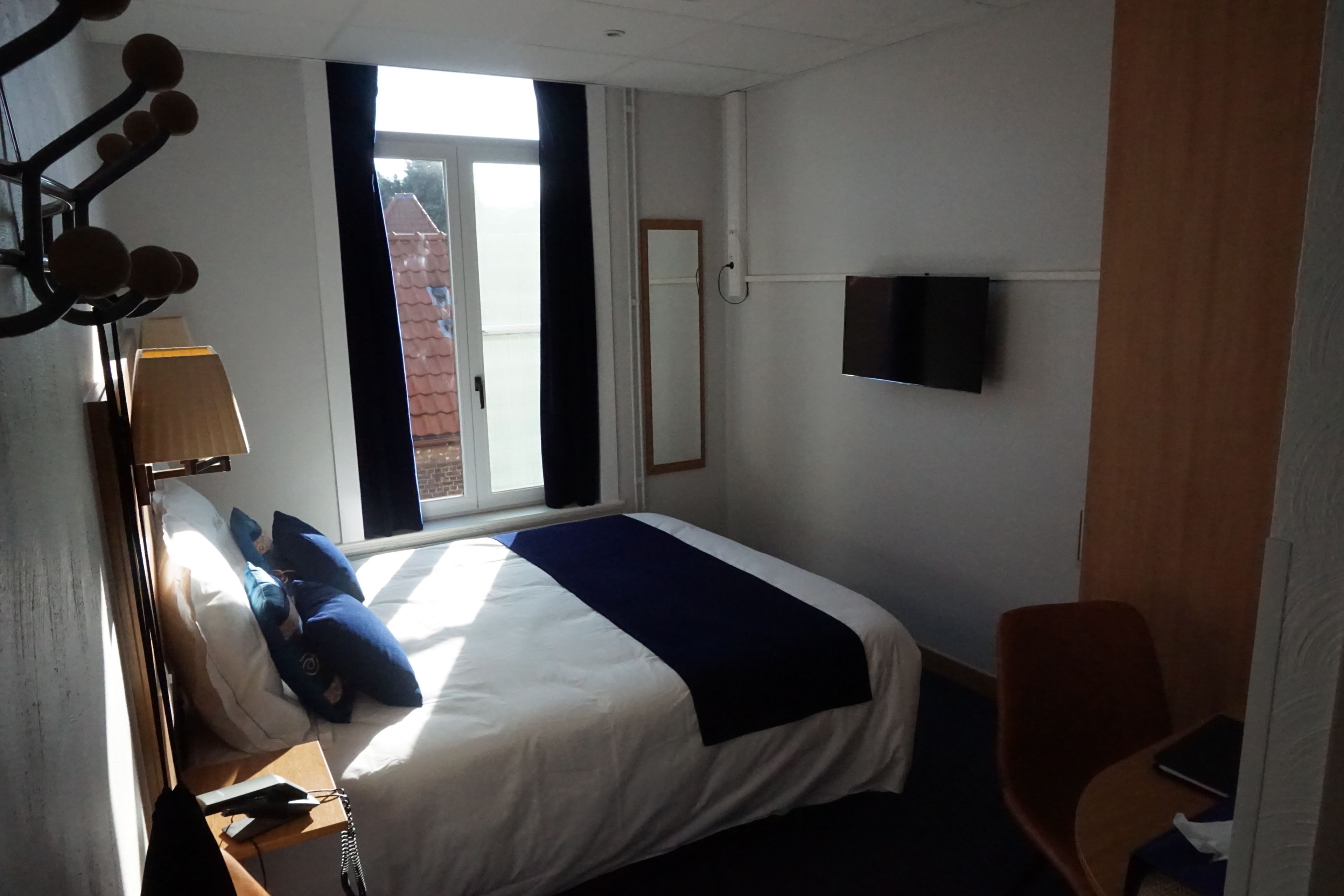 Een hotelkamer met een bed, een bureau met lamp, een stoel, een aan de muur bevestigde televisie en een raam met uitzicht op gebouwen.