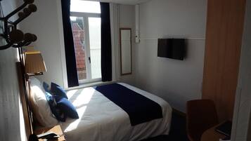 Een hotelkamer met een bed, een bureau met lamp, een stoel, een aan de muur bevestigde televisie en een raam met uitzicht op gebouwen.