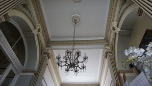 A grand chandelier hanging from a high ceiling with ornate moldings and a large window. - Hotel Asiris (Bruges)