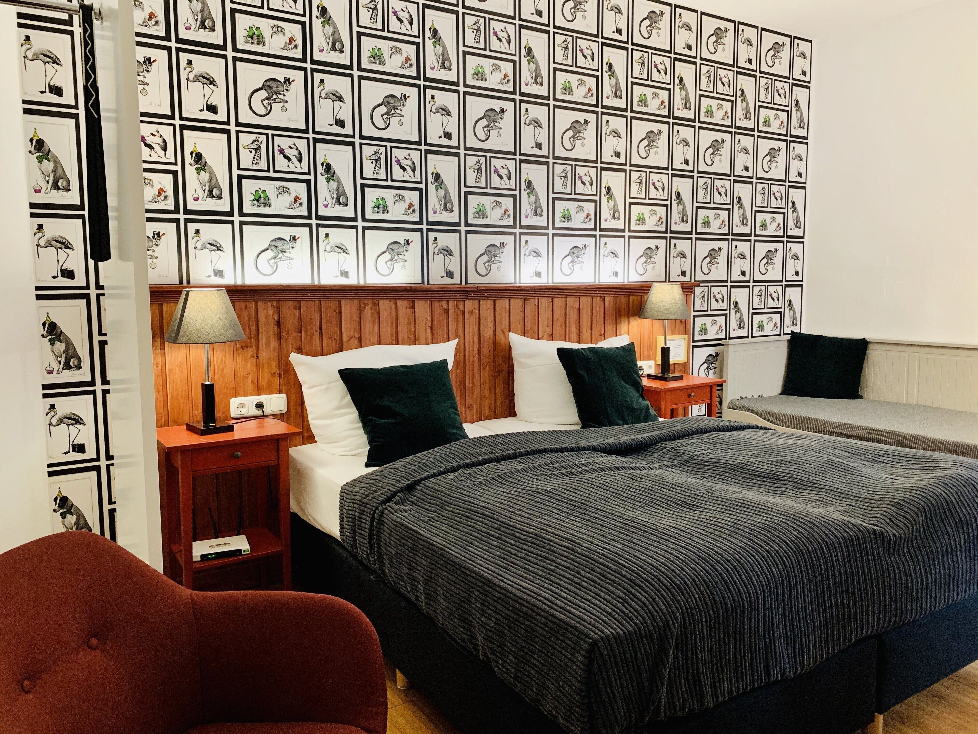 Une chambre d’hôtel avec deux lits, une tête de lit en bois, un fauteuil rouge et un mur décoré d’illustrations d’animaux encadrées.
