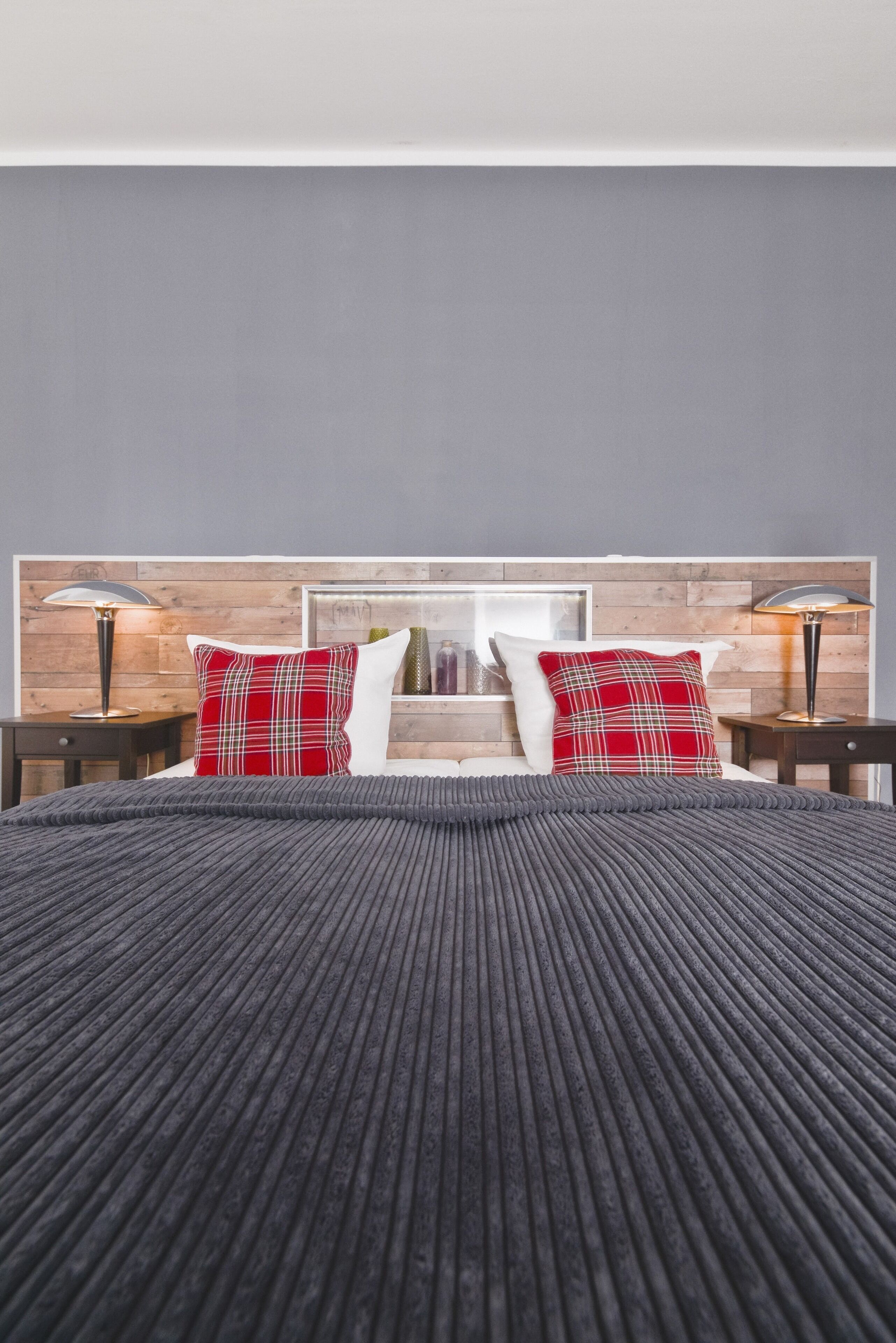 A modern hotel room with a wooden headboard, a large bed with a grey textured bedspread, two red plaid pillows, and two bedside lamps.