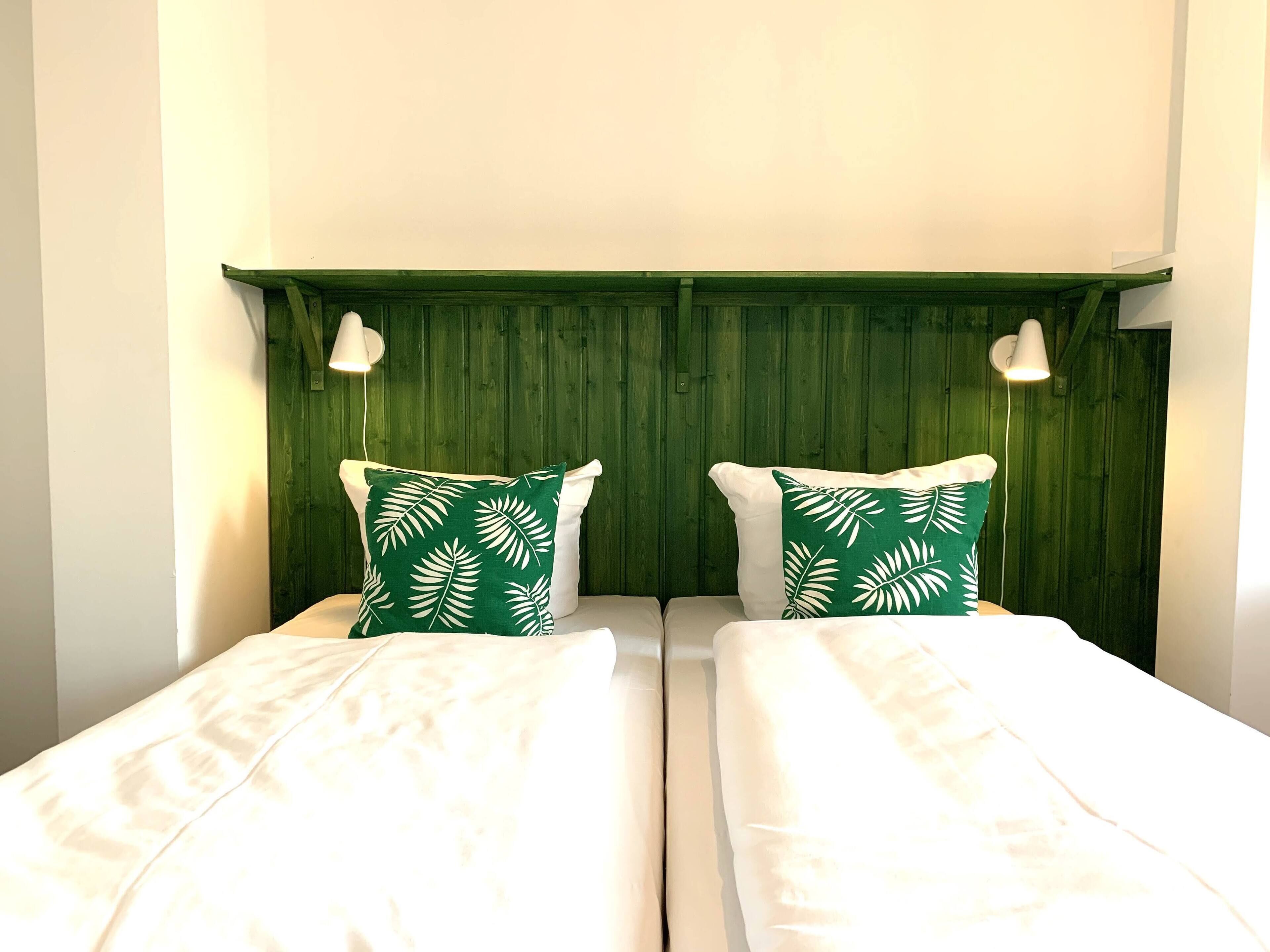 A double bed with white bedding and two green pillows with white leaf patterns, against a green wooden headboard with two wall-mounted lamps.