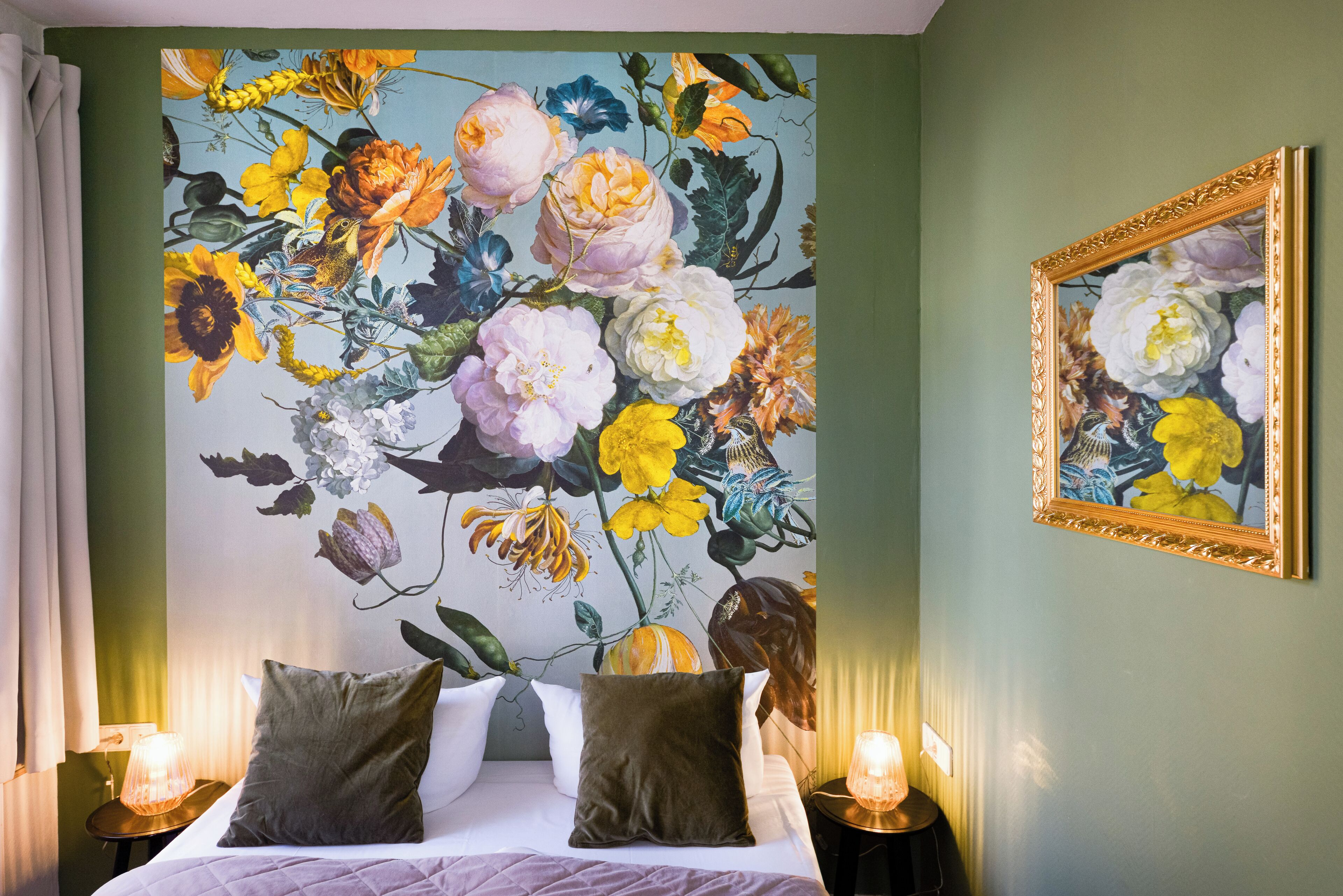A hotel room with a large floral mural on the wall, a bed with multiple pillows, and two bedside lamps.
