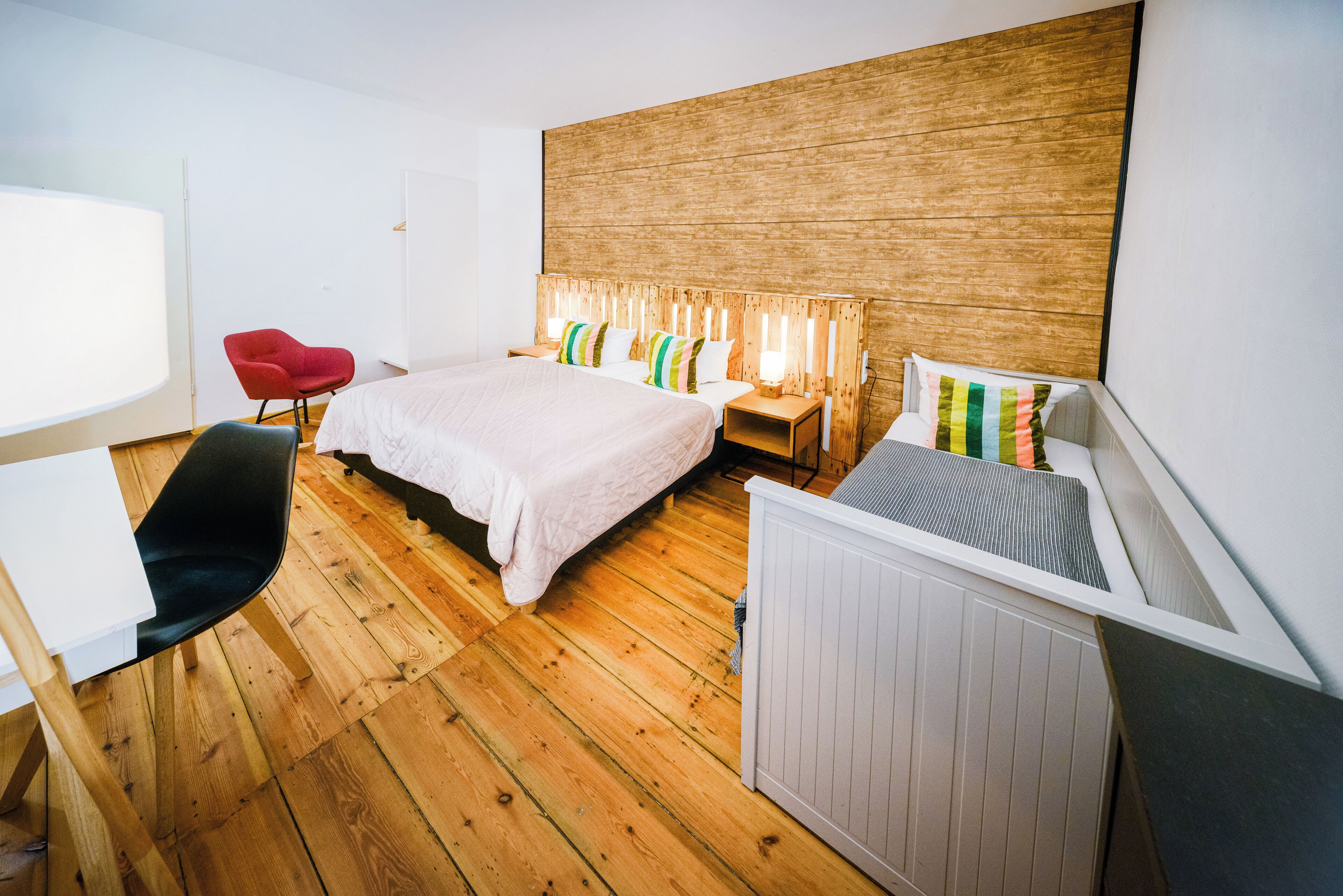 Moderni hotellihuone, jossa on puulattia, valkoisilla tekstiileillä varustettu sänky, punainen tuoli, työpöytä ja patteri.