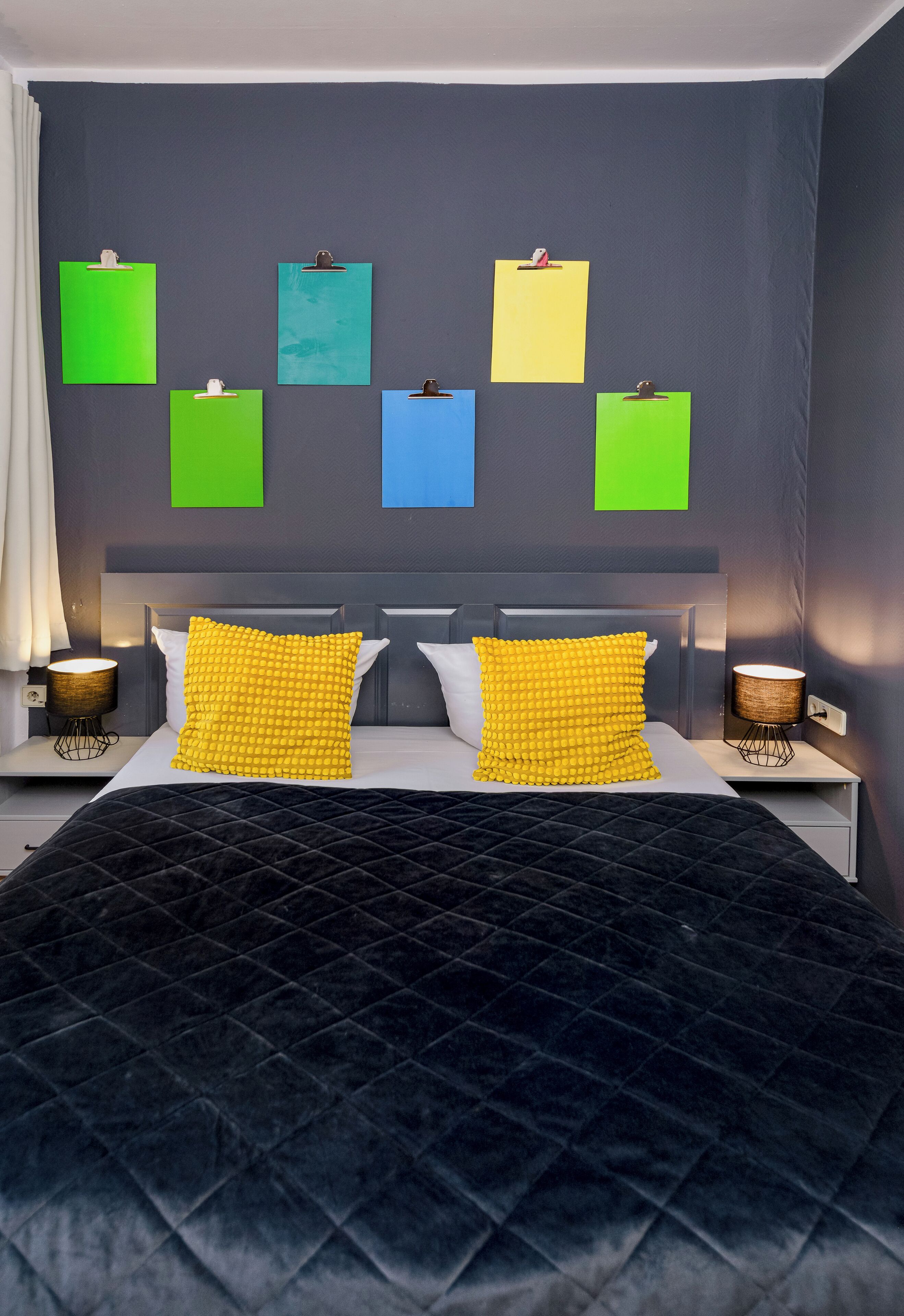 A bedroom with a dark blue quilted bedspread, two yellow pillows, and a grey pillow. There are four clipboards on the wall, and two bedside lamps.