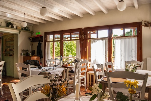 A dining area with white chairs and tables, large windows with curtains, and hanging light fixtures.