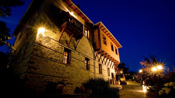 A stone building with a balcony and multiple windows, illuminated by warm lights at night. - Viraggas (Polygyros)