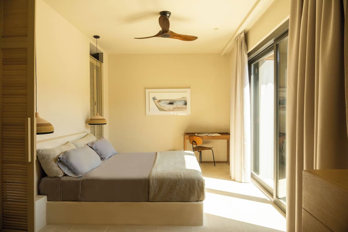 Une chambre à coucher comprenant un lit, un bureau, une chaise, un ventilateur de plafond et une fenêtre avec des rideaux.