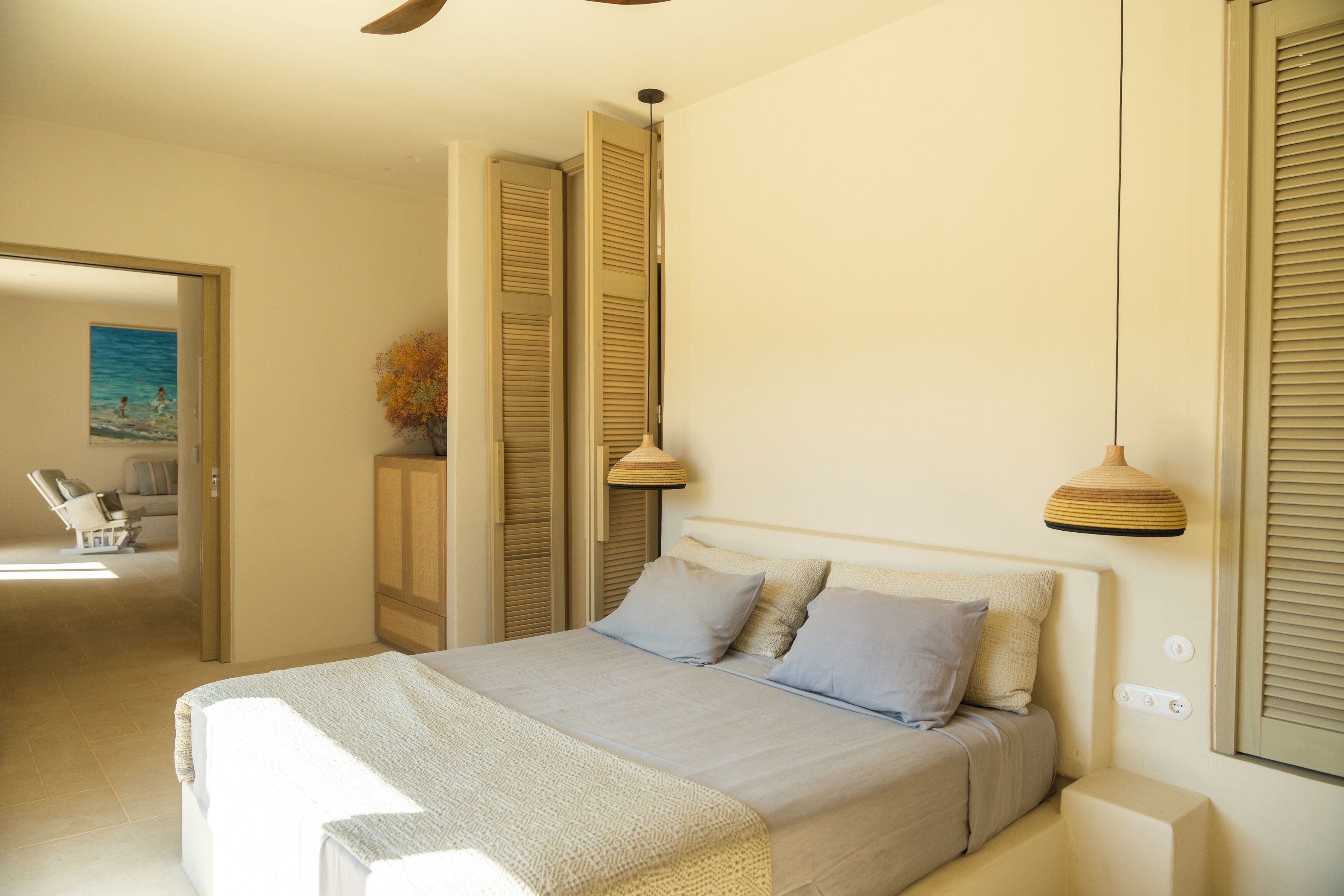 Quarto com cama, abajures e vista para uma praia através de uma porta aberta.