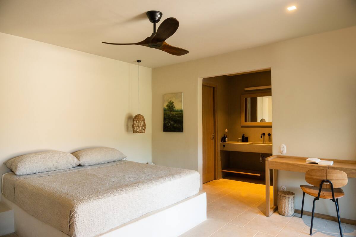 Une chambre à coucher comprenant un lit, un bureau, une chaise, un ventilateur de plafond et une salle de bain visible à travers une porte ouverte.