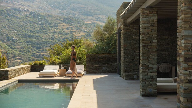 A pool area with a stone wall, a lounge chair, and a person walking. - Onar Andros (Andros)