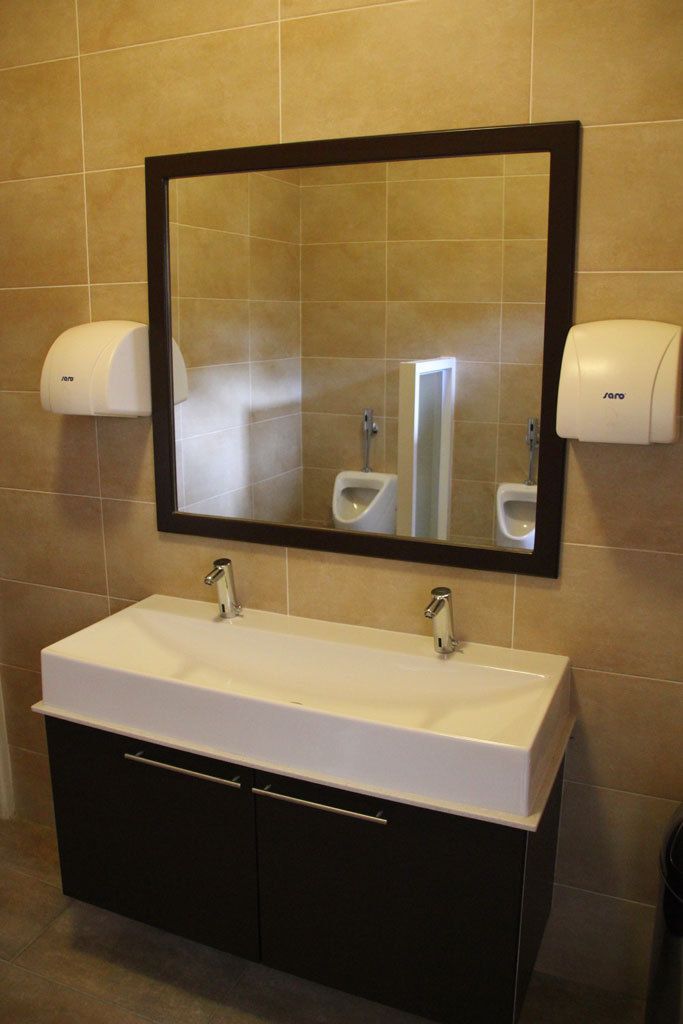 Un baño con un lavamanos blanco, dos grifos, un espejo grande y dos dispensadores de toallas de papel.