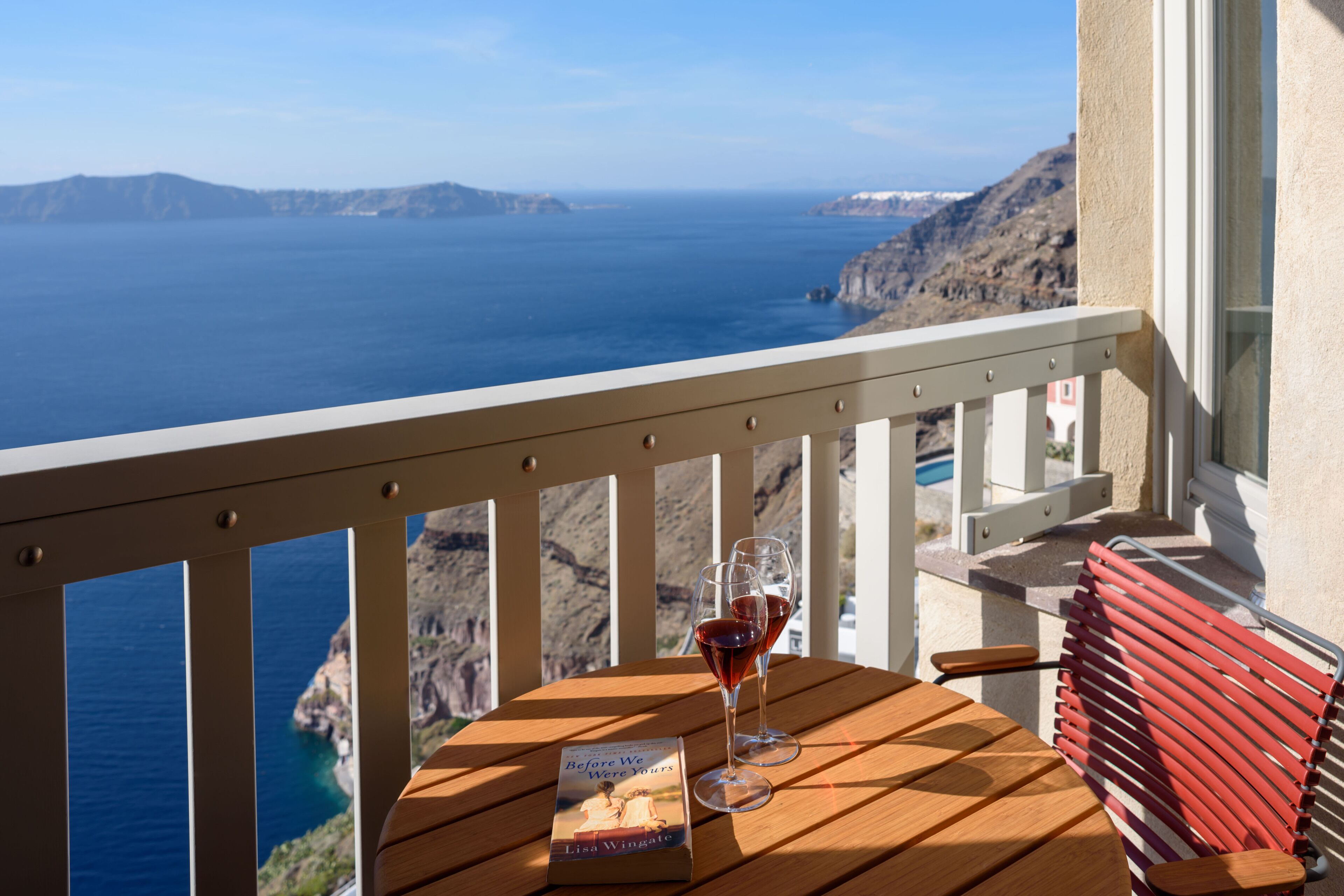 Una mesa de madera con un libro titulado "Before We Were Yours" de Lisa Wingate, dos copas de vino con vino tinto y un banco rojo en un balcón con vistas a la costa.
