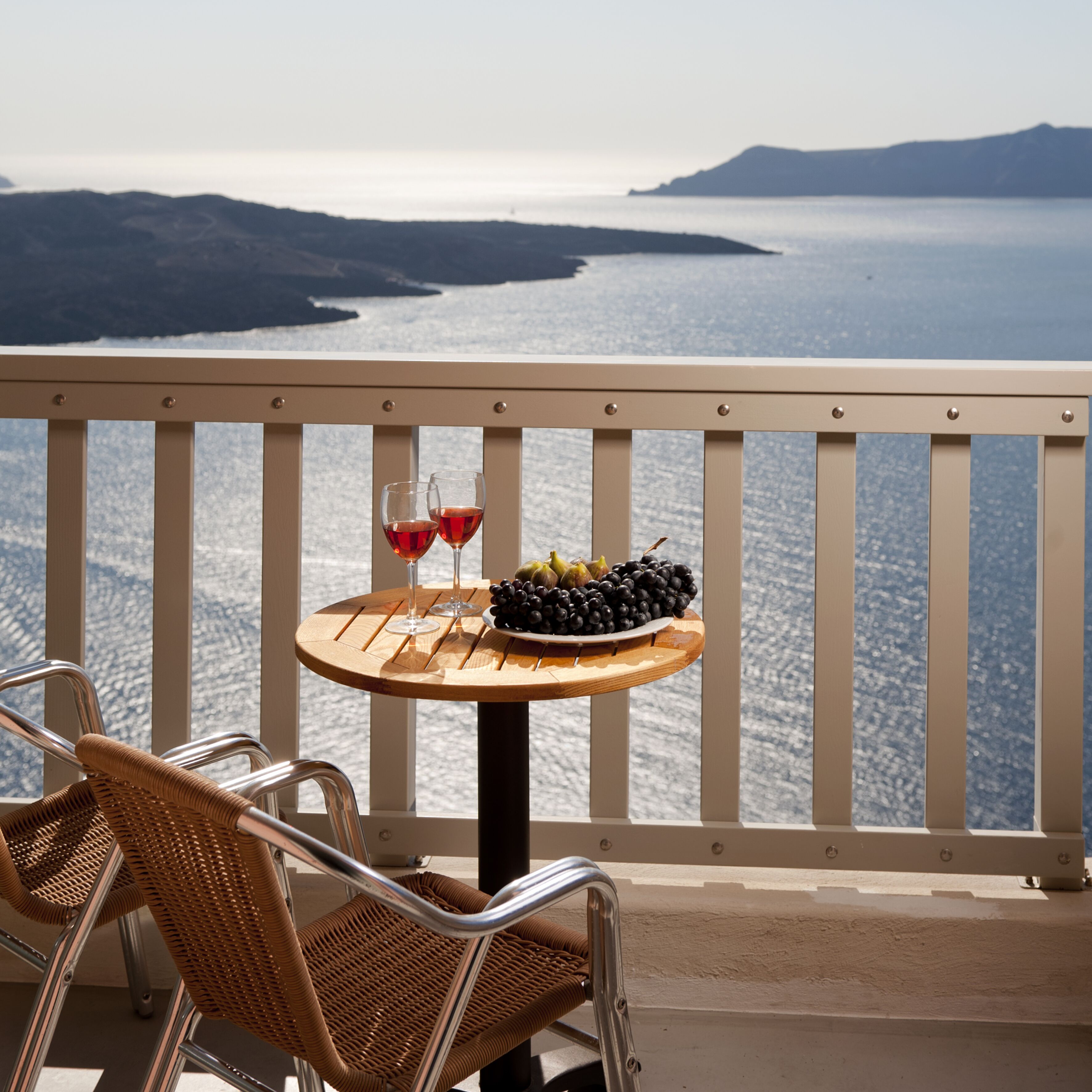 A balcony with a table set for two, featuring a plate of grapes and two glasses of red wine.