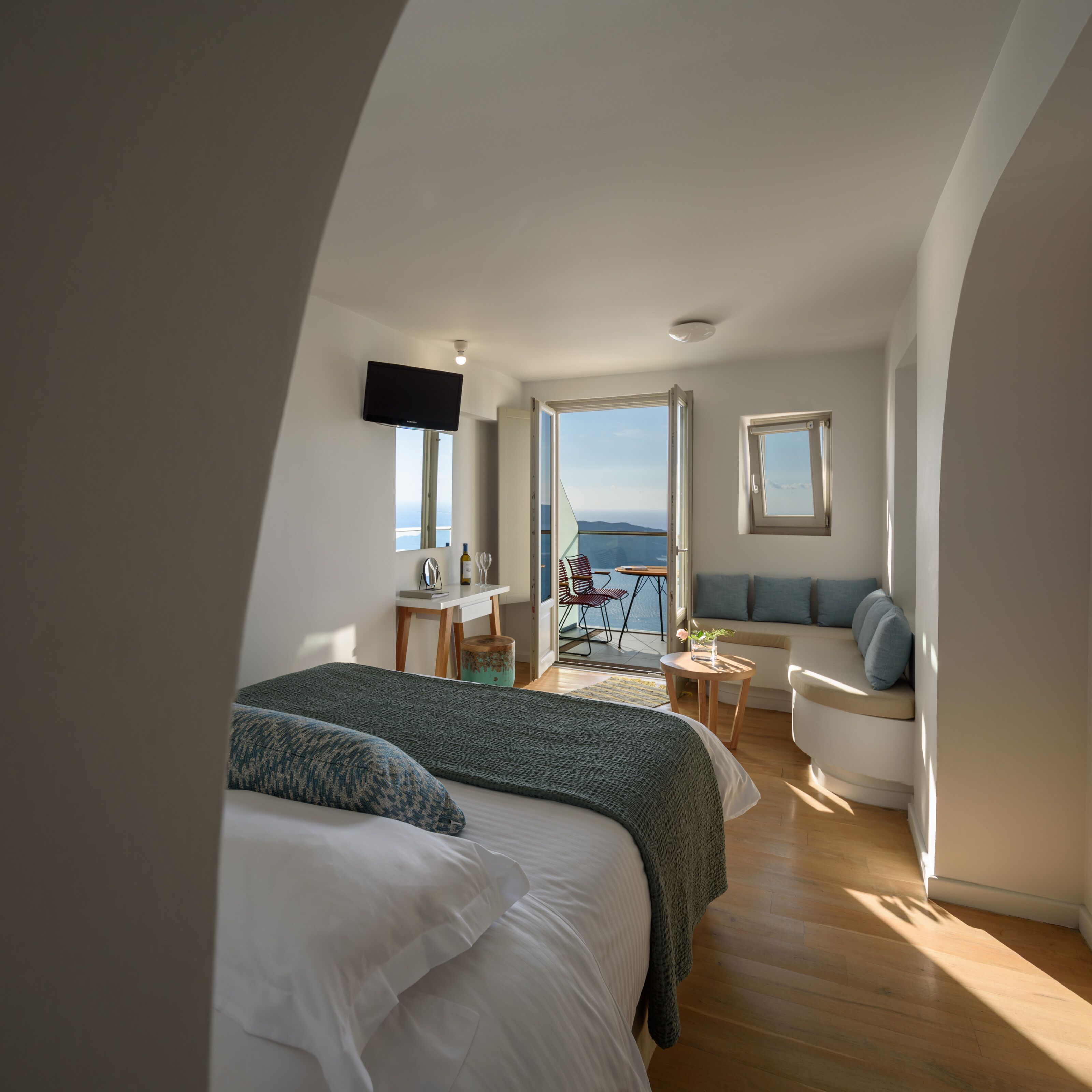 A modern bedroom with a large bed, a desk, a chair, and a sofa. There is a window with a view of the sea.