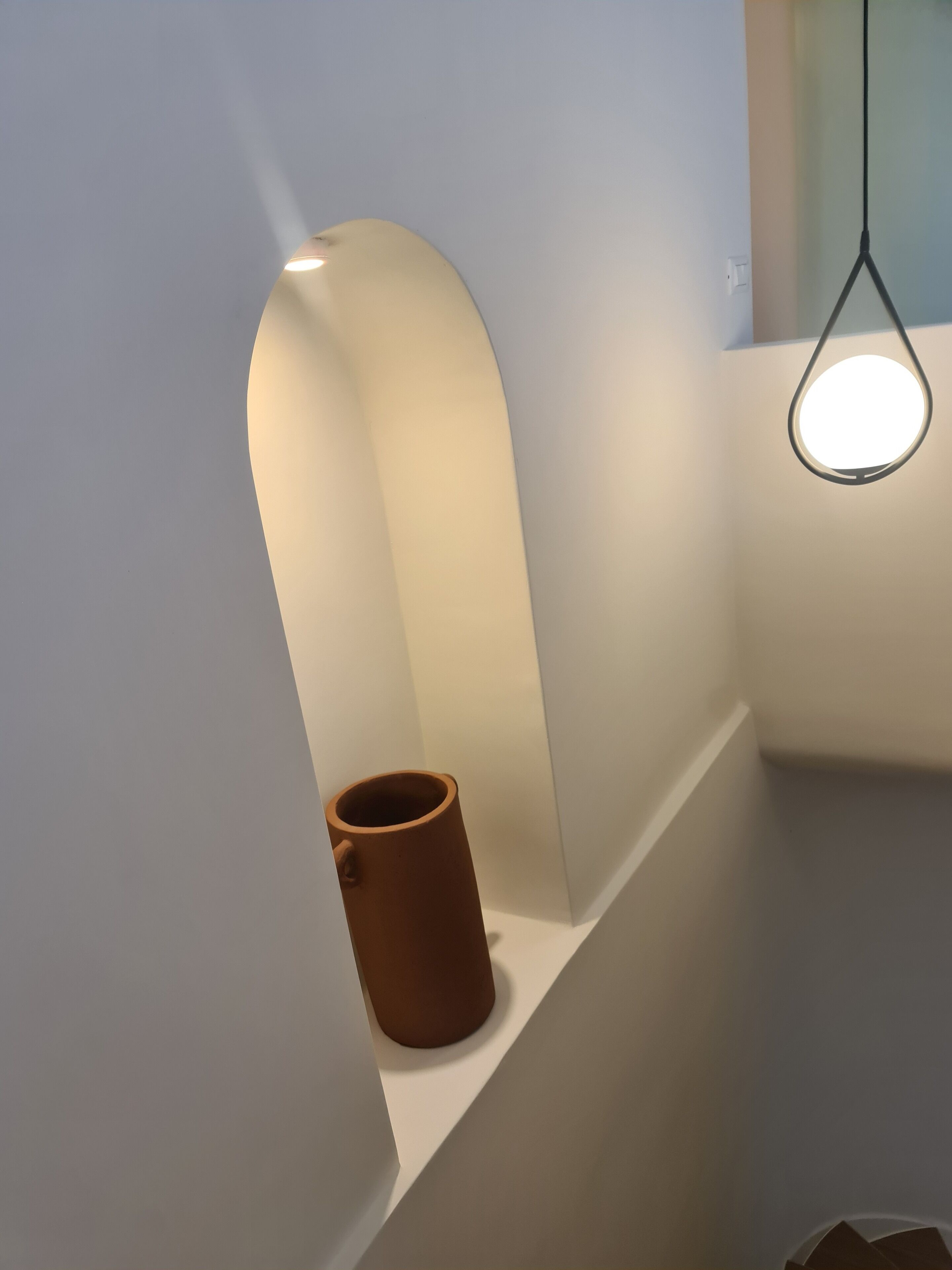 Un angolo di una stanza con una parete bianca, una pentola di ceramica marrone su uno scaffale e una lampada a sospensione.