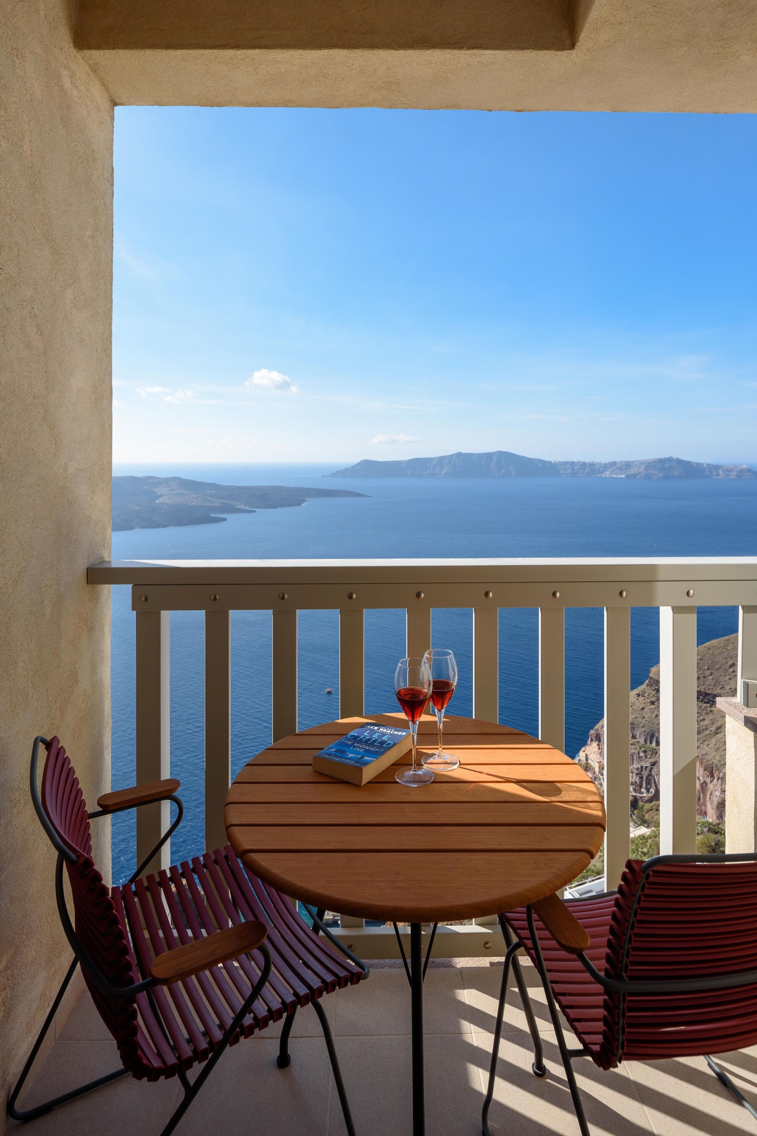 Uma varanda com mesa de madeira e duas cadeiras, oferecendo uma vista panorâmica do mar e montanhas ao longe.