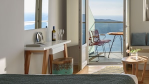 Un dormitorio con cama, escritorio, silla, una mesita con un florero y balcón con vistas.