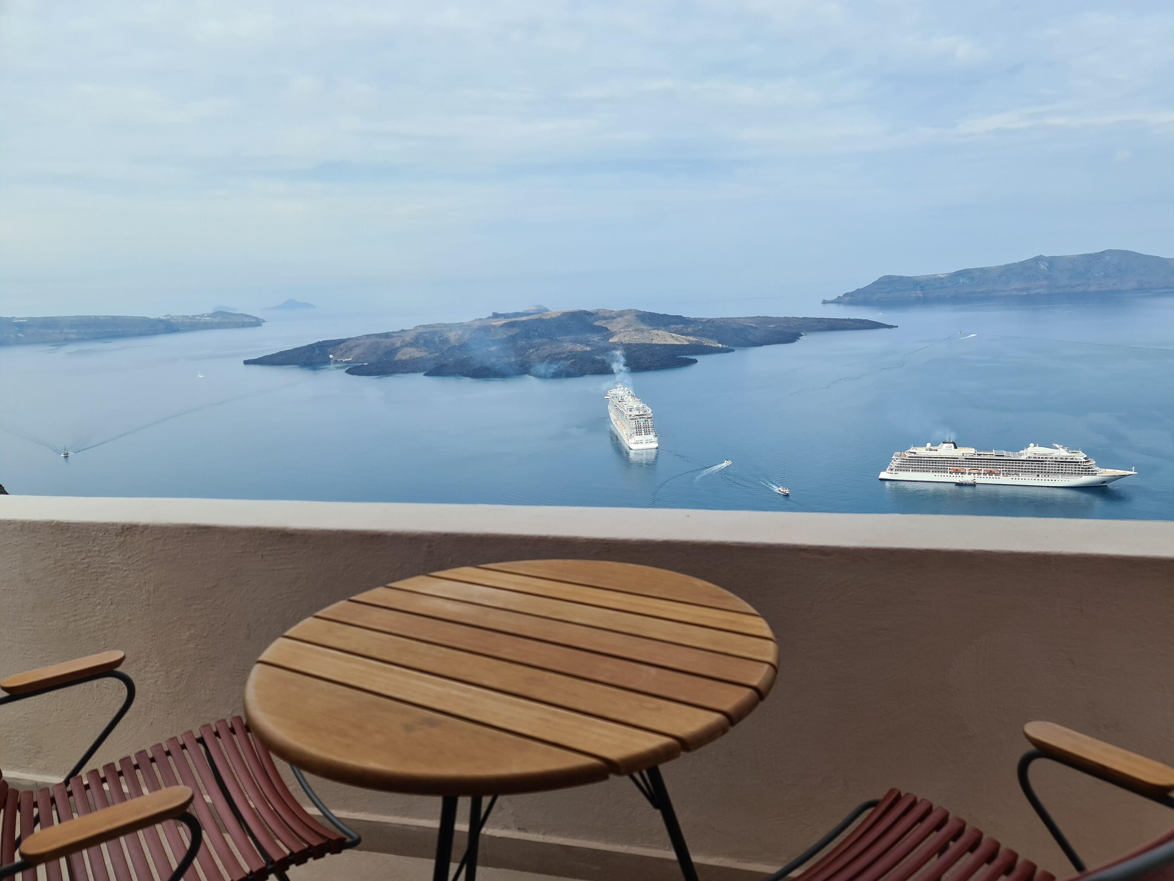Un balcon avec une table et des chaises donnant sur une mer calme, où l’on peut apercevoir deux navires de croisière.