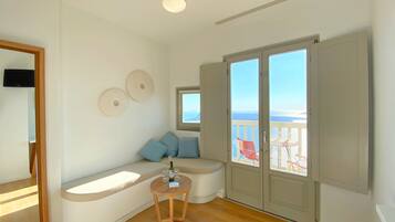 Ein Zimmer mit einer gebogenen Sitzbank, einem kleinen Tisch mit Gläsern und einer Flasche sowie einer Glastür zum Balkon mit Meerblick.